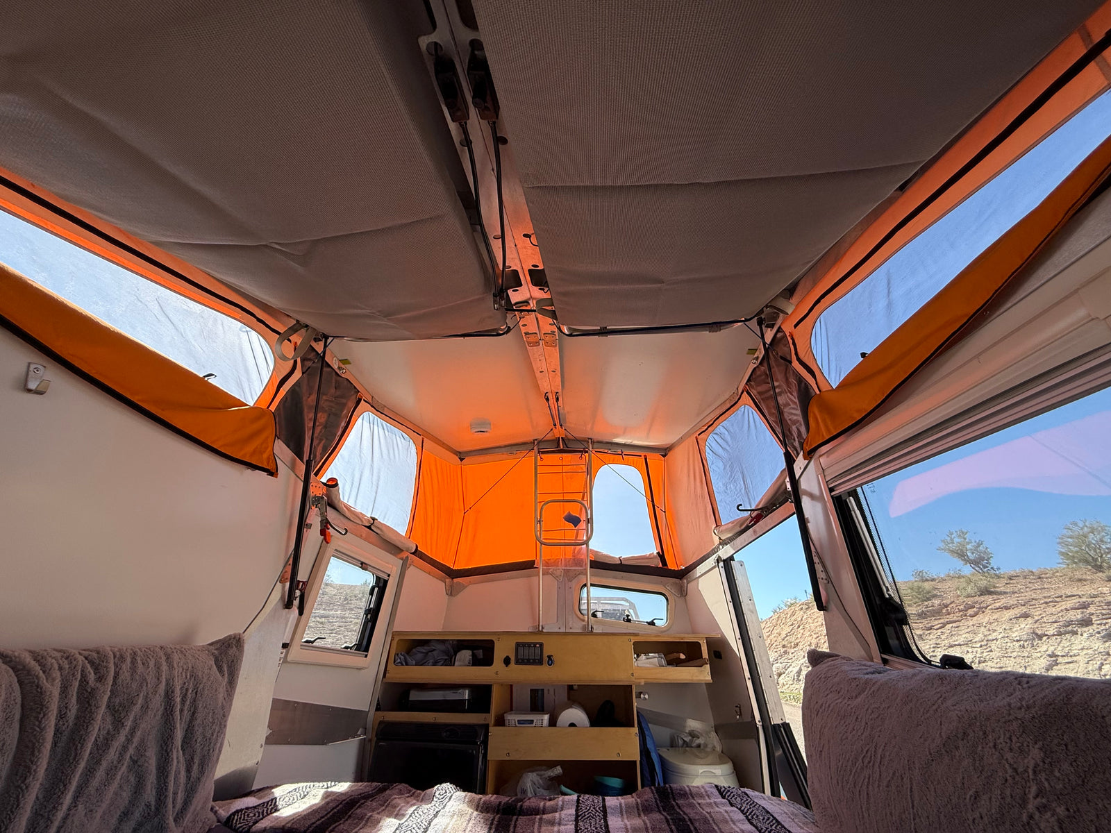 Inside a pop-up rv rental: compact interior with bed, small kitchen, and panoramic windows showing a desert view.