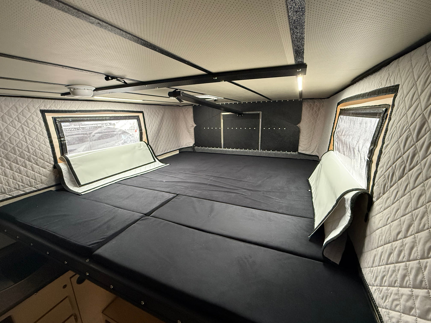 Inside an RV rental: a cozy sleeping area featuring a segmented black mattress, quilted walls, and reflective window covers.