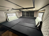 Inside an RV rental: a cozy sleeping area featuring a segmented black mattress, quilted walls, and reflective window covers.