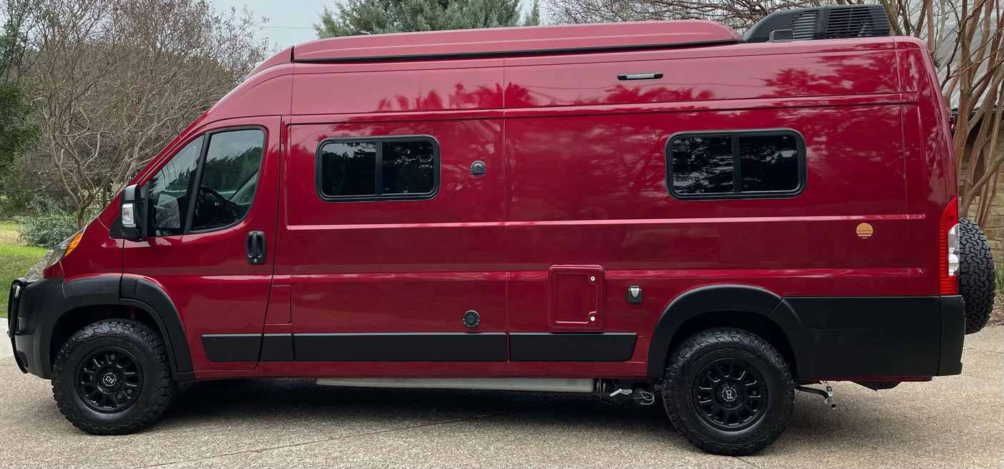 This robust, dark red RV rental camper van is ready for adventure. Side view shows off-road tires, custom windows & rooftop unit.