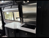 Modern RV rental kitchen with white speckled countertop, induction cooktop, and faucet, featuring a snowy view.