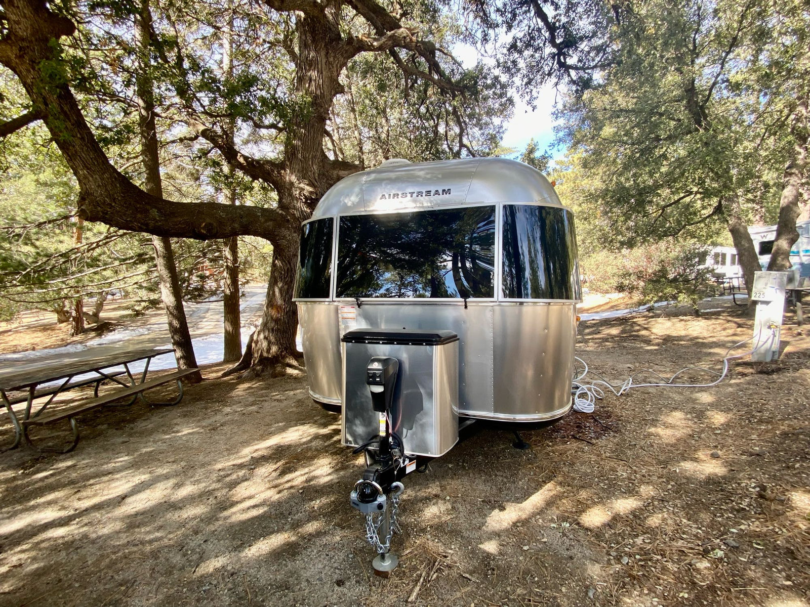 This Airstream travel trailer, likely an RV rental, is perfectly parked at a scenic dirt campsite under large shade trees.