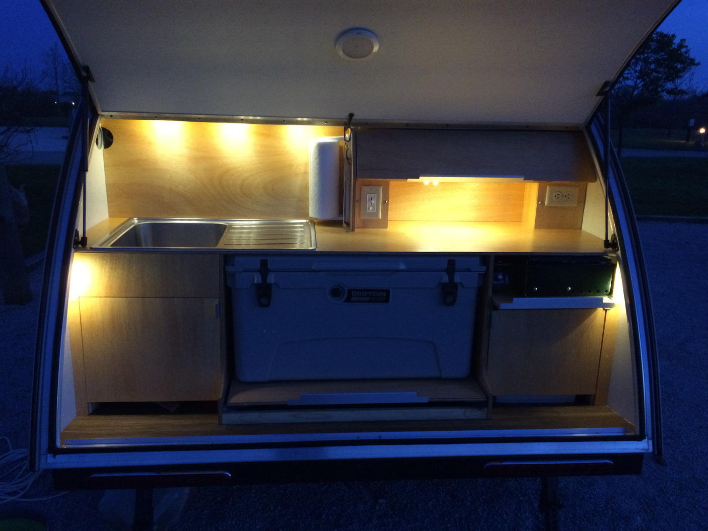 Detailed view of an RV rental's illuminated rear galley, featuring a sink, storage compartments, and a large cooler.