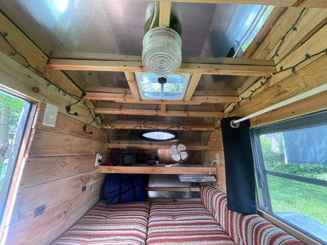 Inside an RV rental, showing a cozy bed, wooden walls, roof vent, and a shelf with a fan/heater.