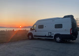 Experience epic views in your RV rental! White Winnebago Solis camper van parked by the water at sunset with mountains.