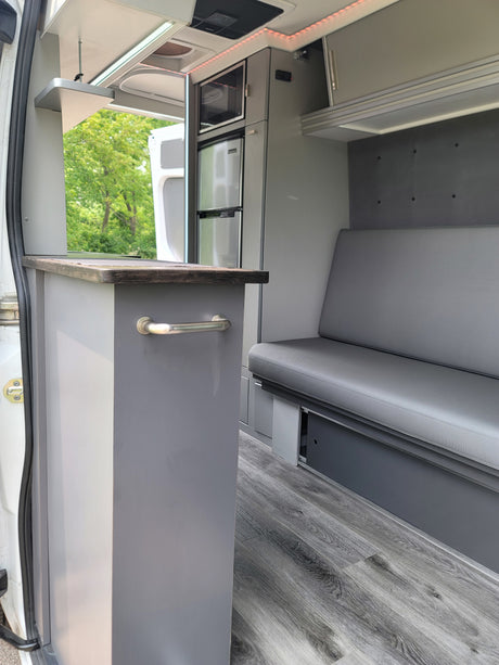 Inside a modern RV rental: grey kitchen module with microwave, fridge, and matching bench seating. Wood-look flooring.