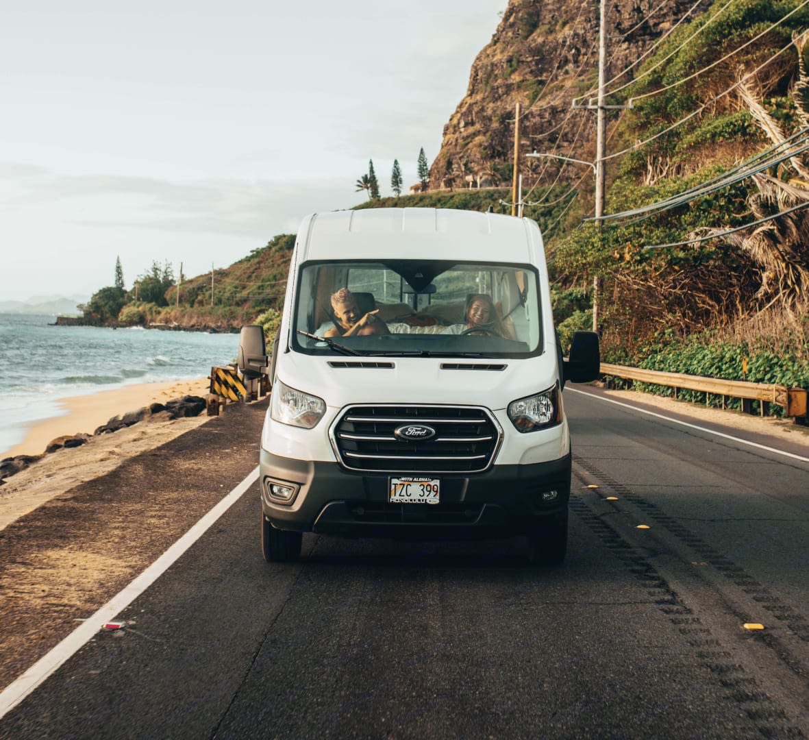 Two people enjoy a Hawaiian coastal road trip in their white rv rental van, with ocean and mountains in the background.