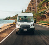 Two people enjoy a Hawaiian coastal road trip in their white rv rental van, with ocean and mountains in the background.