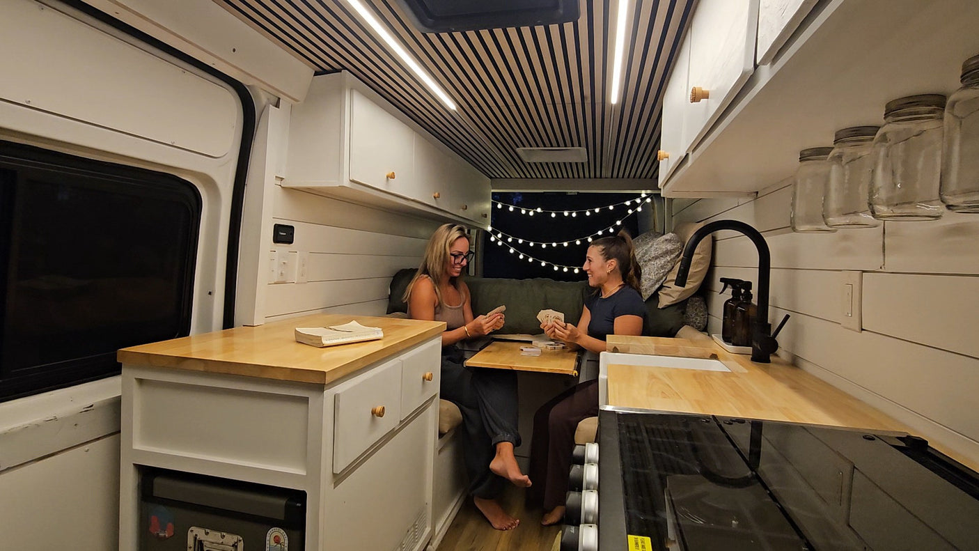 Two women enjoy cards inside a stylish RV rental camper van, featuring a modern, cozy interior with wooden accents and string lights.