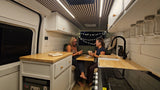 Two women enjoy cards inside a stylish RV rental camper van, featuring a modern, cozy interior with wooden accents and string lights.