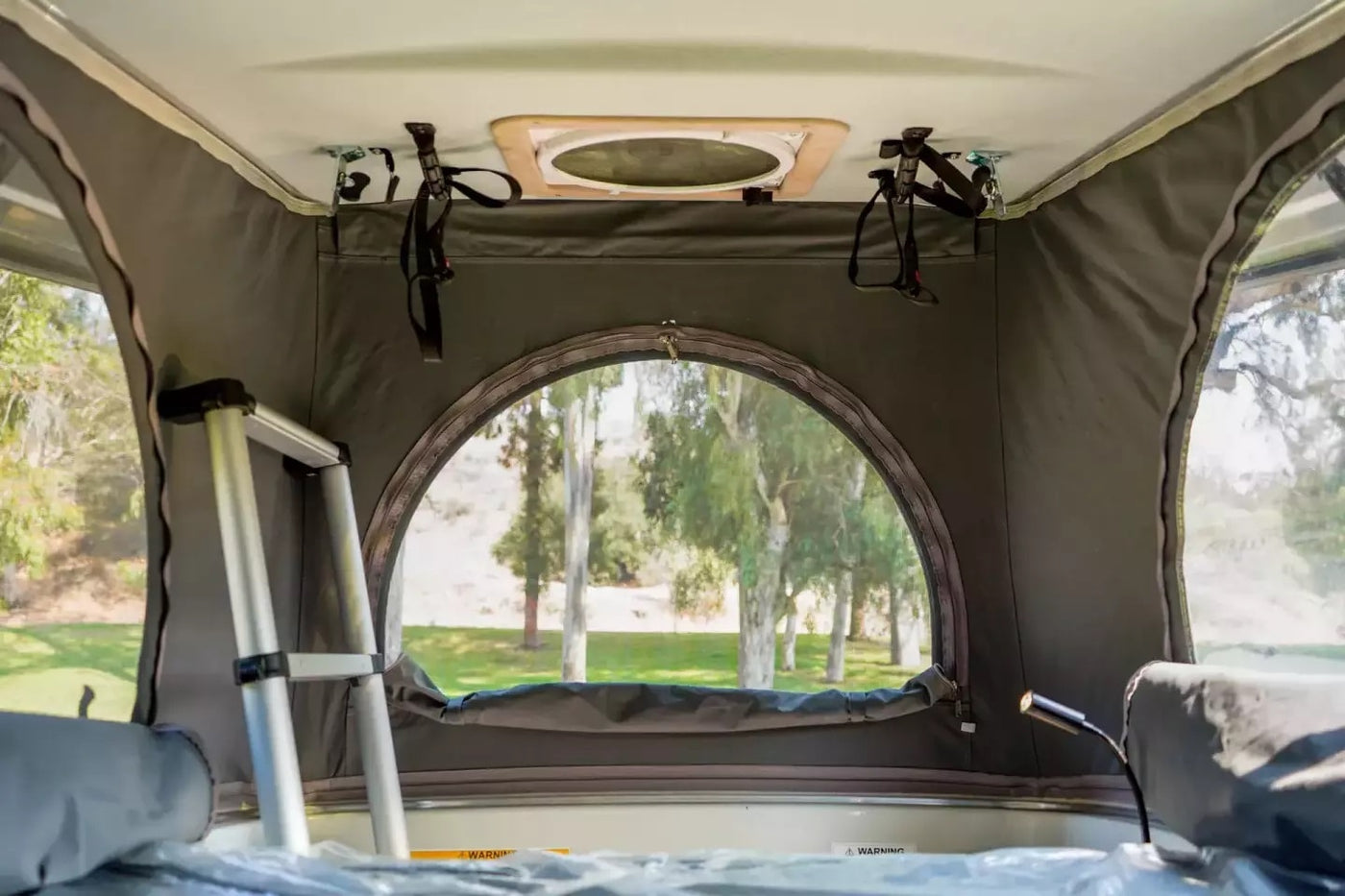 Peek inside an RV rental's cozy pop-top tent with a round window view of lush trees & grass. Ladder visible. — photo 8