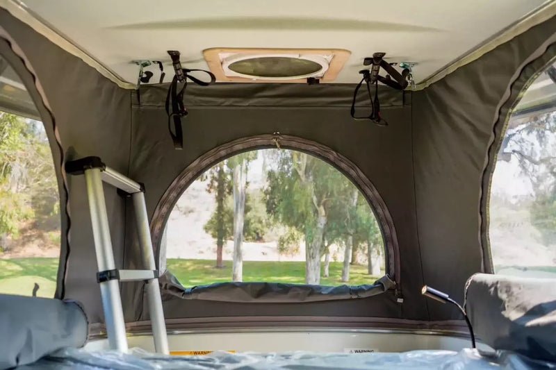 Peek inside an RV rental's cozy pop-top tent with a round window view of lush trees & grass. Ladder visible. — photo 8