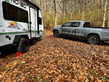 A grey pickup truck and white travel trailer ('No Boundaries' RV) parked in a leaf-covered forest campsite, ideal for rv rental.
