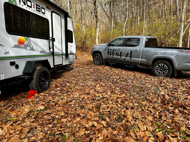 A grey pickup truck and white travel trailer ('No Boundaries' RV) parked in a leaf-covered forest campsite, ideal for rv rental. — photo 1