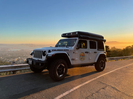 White Jeep Rubicon with rooftop tent, perfect for an overland RV rental adventure, overlooking a hazy city at sunset.