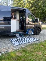RV rental setup: A woman in a dark grey camper van with an open door, two chairs & a mat outside on gravel at a campsite.