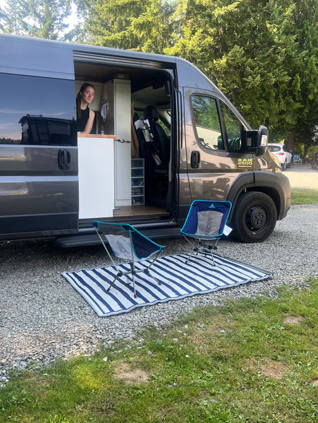 RV rental setup: A woman in a dark grey camper van with an open door, two chairs & a mat outside on gravel at a campsite.