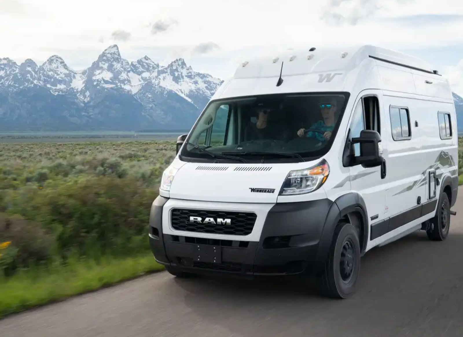 White Winnebago RV rental, driven by a person in blue sunglasses, on a scenic road with snow-capped mountains.