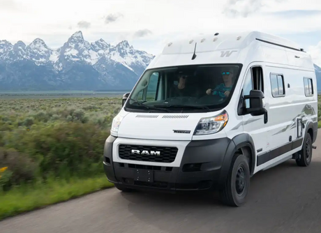 White Winnebago RV rental, driven by a person in blue sunglasses, on a scenic road with snow-capped mountains.