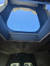 Looking up through the open pop-top roof of this RV rental camper van, showcasing a cozy wood-paneled interior and blue sky.