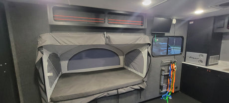 RV rental interior: grey fold-down bunk, overhead storage with red bungees, TV, and compact kitchen with 'Flyer' appliance.