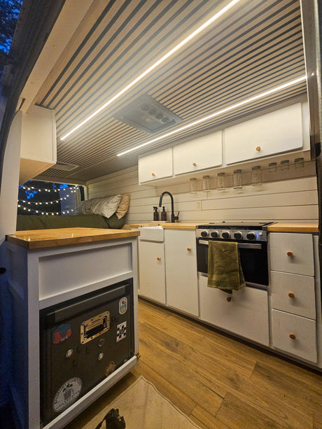 Inside a modern RV rental: a compact kitchen with a stove, sink, and cabinets, plus a cozy sleeping area, all brightly lit.