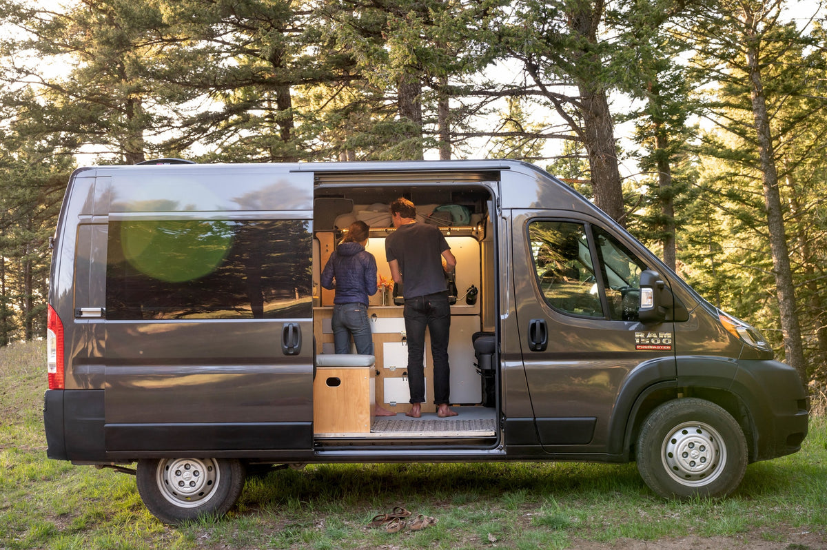 This custom Ram Promaster camper van rv rental offers outdoor living. Two people cook in its cozy interior amidst nature. — photo 12