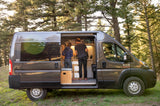 This custom Ram Promaster camper van rv rental offers outdoor living. Two people cook in its cozy interior amidst nature.