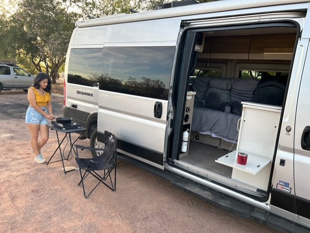 This RV rental offers outdoor cooking & cozy van life! A woman cooks outside her silver camper van, door open to show the bed.