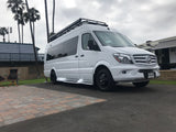 This white Mercedes Sprinter RV rental, an adventure van with a roof rack, is parked on a paved site near palm trees.