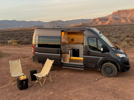 An RV rental camper van, side door open to its lit interior, with chairs and a fire pit, parked in a desert landscape.