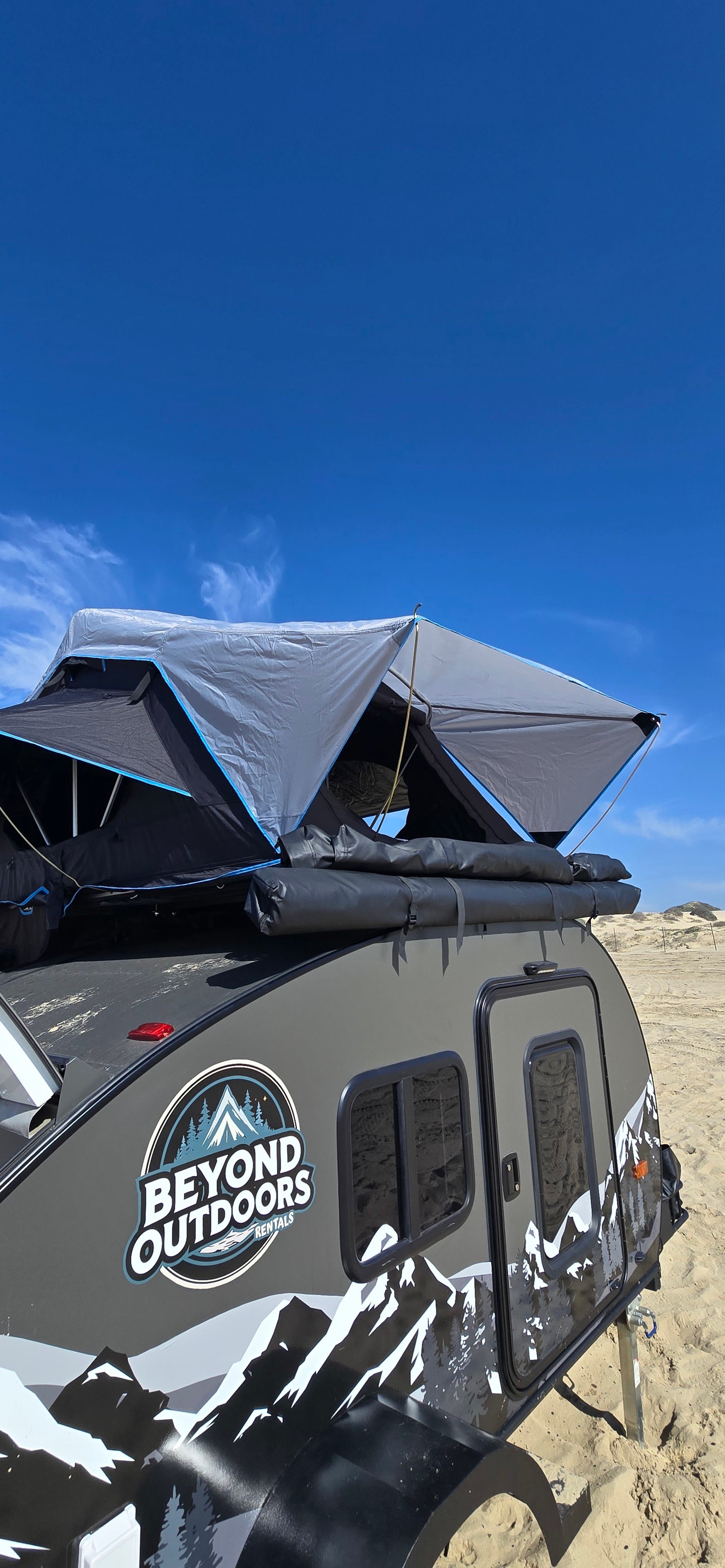 Beyond Outdoors rv rental teardrop camper with a rooftop tent deployed on sand under a clear blue sky.
