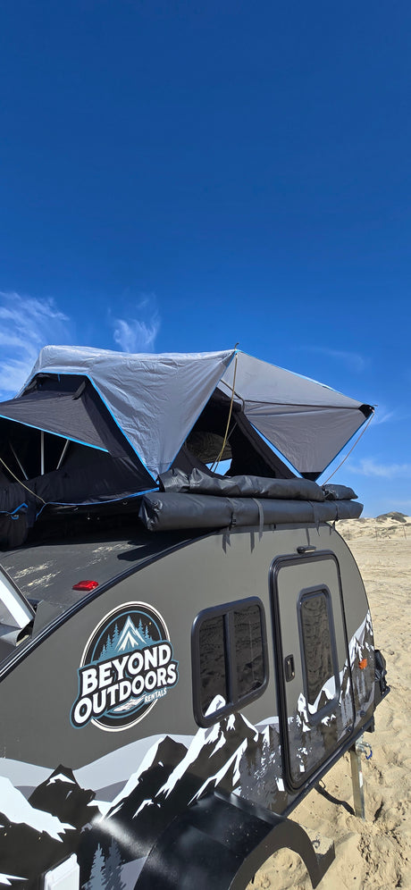 Beyond Outdoors rv rental teardrop camper with a rooftop tent deployed on sand under a clear blue sky.