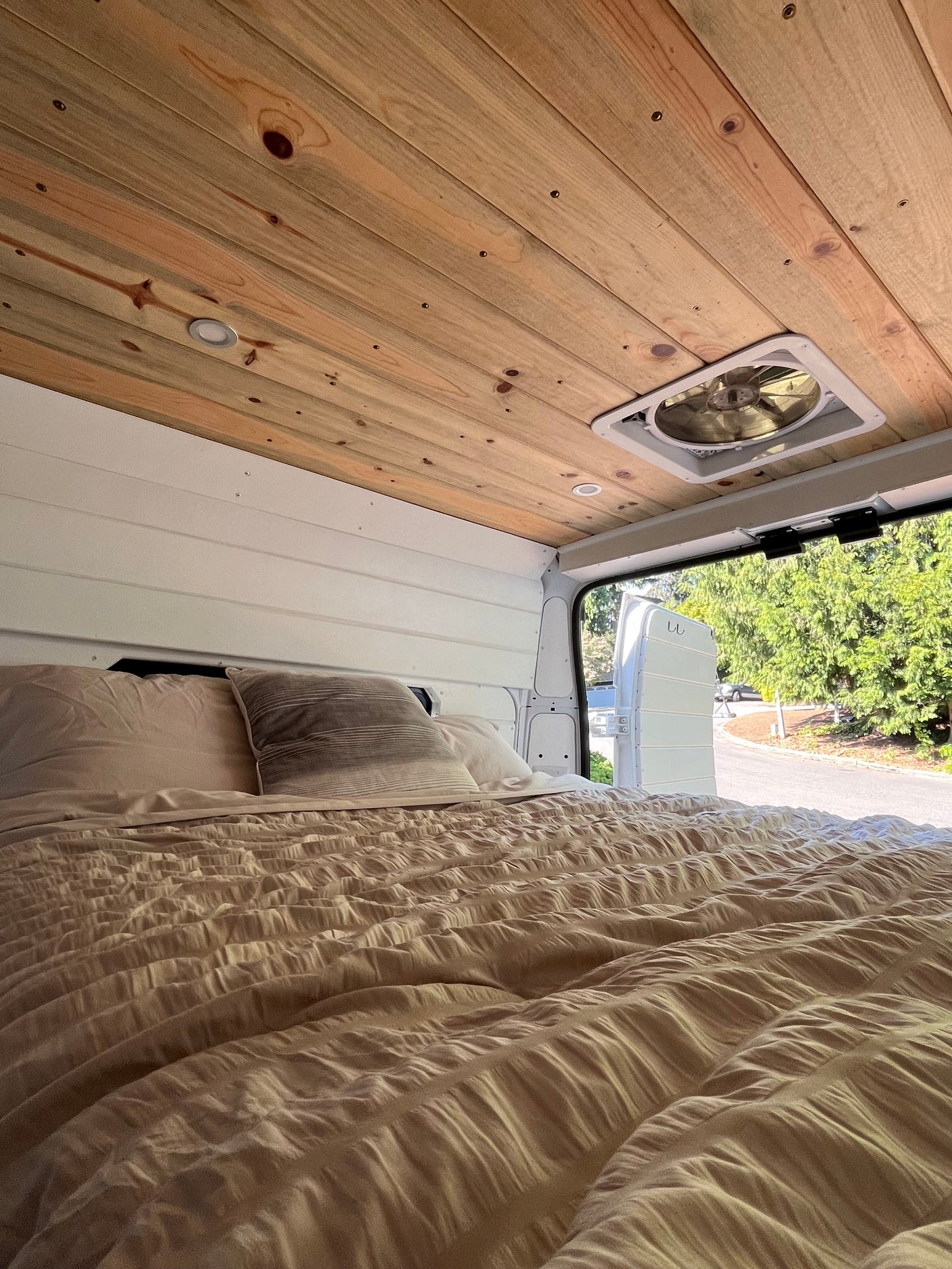 Cozy RV rental bedroom with wood ceiling, fan, soft bed, and open back doors revealing a tree-lined street outside.