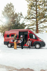 Snowy RV rental trip: Man with snowboard, woman by red camper van, chair, blanket in winter scene.
