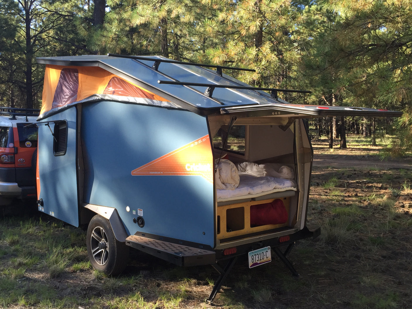 This blue pop-top camper trailer, perfect for an rv rental, is ready for forest adventures with its open rear revealing a cozy bed.