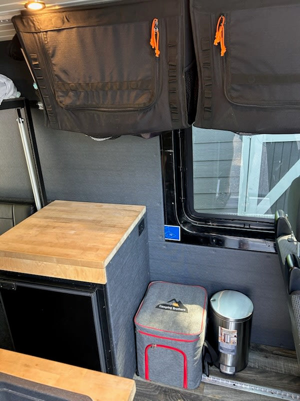 This RV rental interior features practical overhead storage, a mini-fridge with counter, cooler, and trash bin.