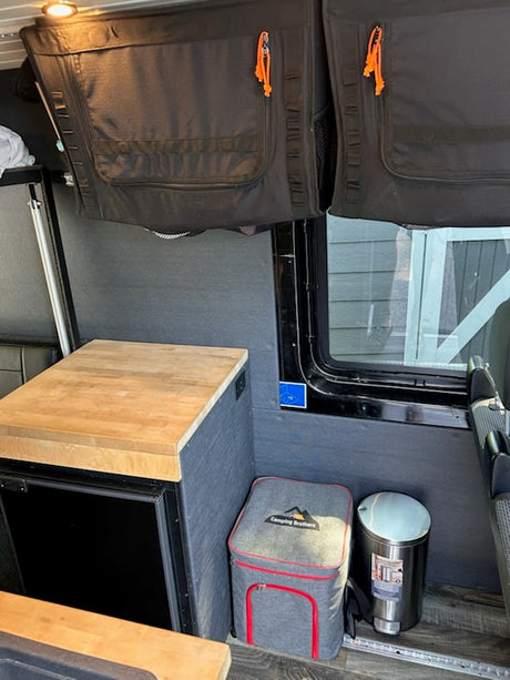 This RV rental interior features practical overhead storage, a mini-fridge with counter, cooler, and trash bin.