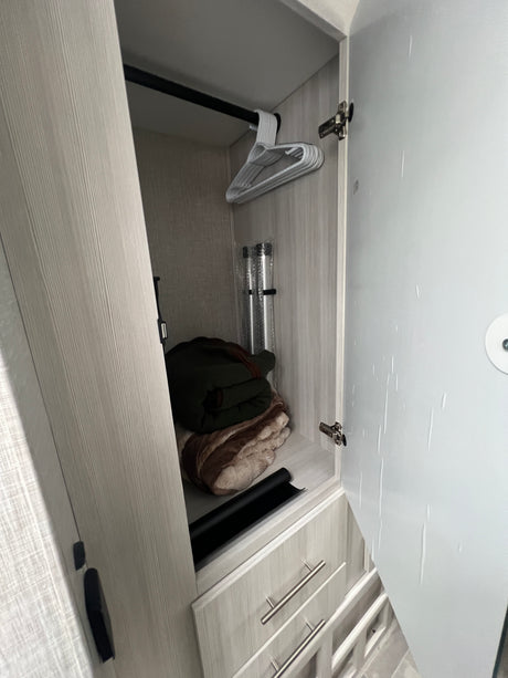 Inside an RV rental, an open closet reveals a hanging rod with hangers, rolled blankets, and two cylindrical items. Drawers are visible below.