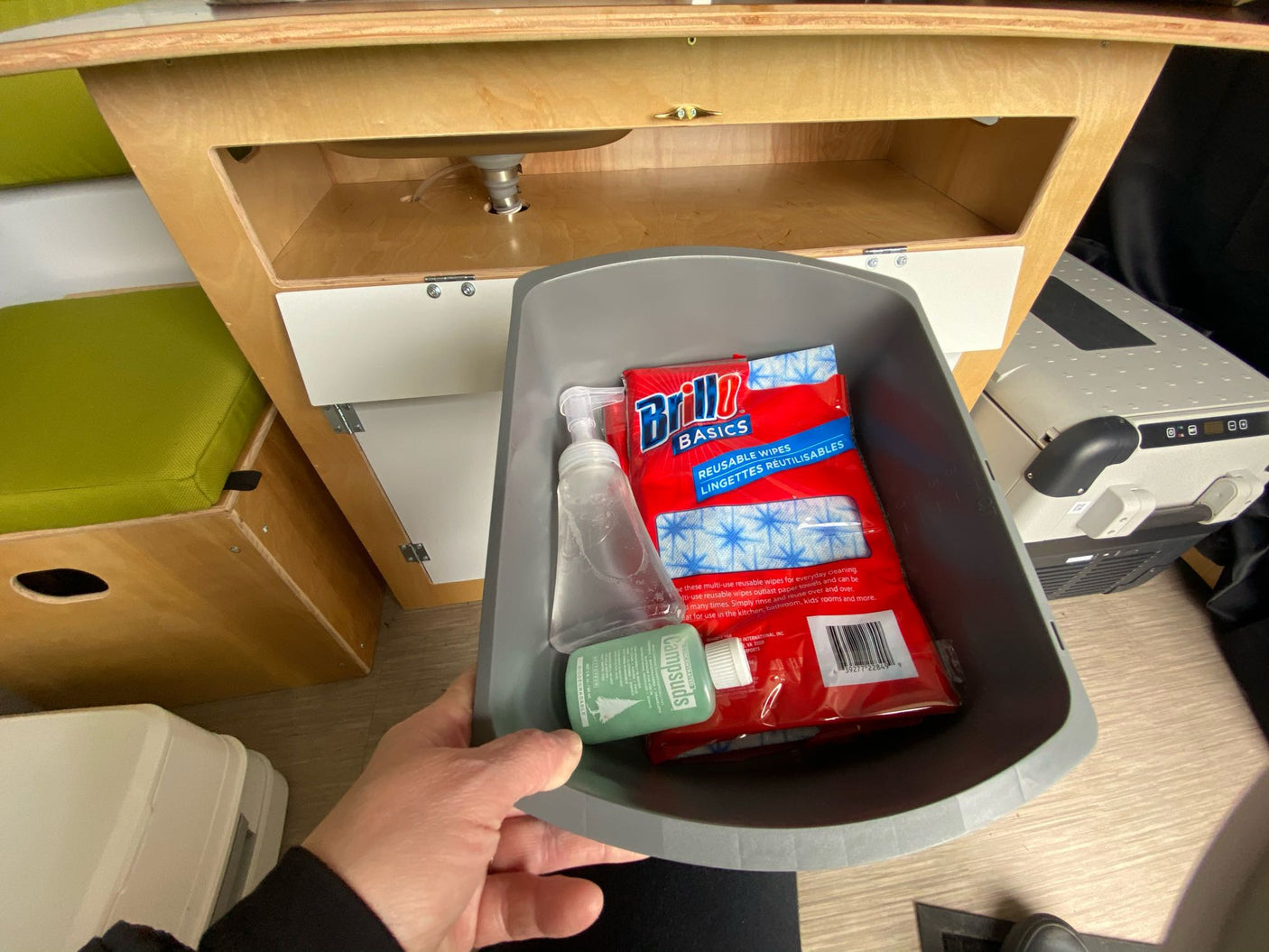 An RV rental kitchen area features cleaning supplies: Brillo wipes, Campsuds, and soap in a bin, ready by the portable sink.