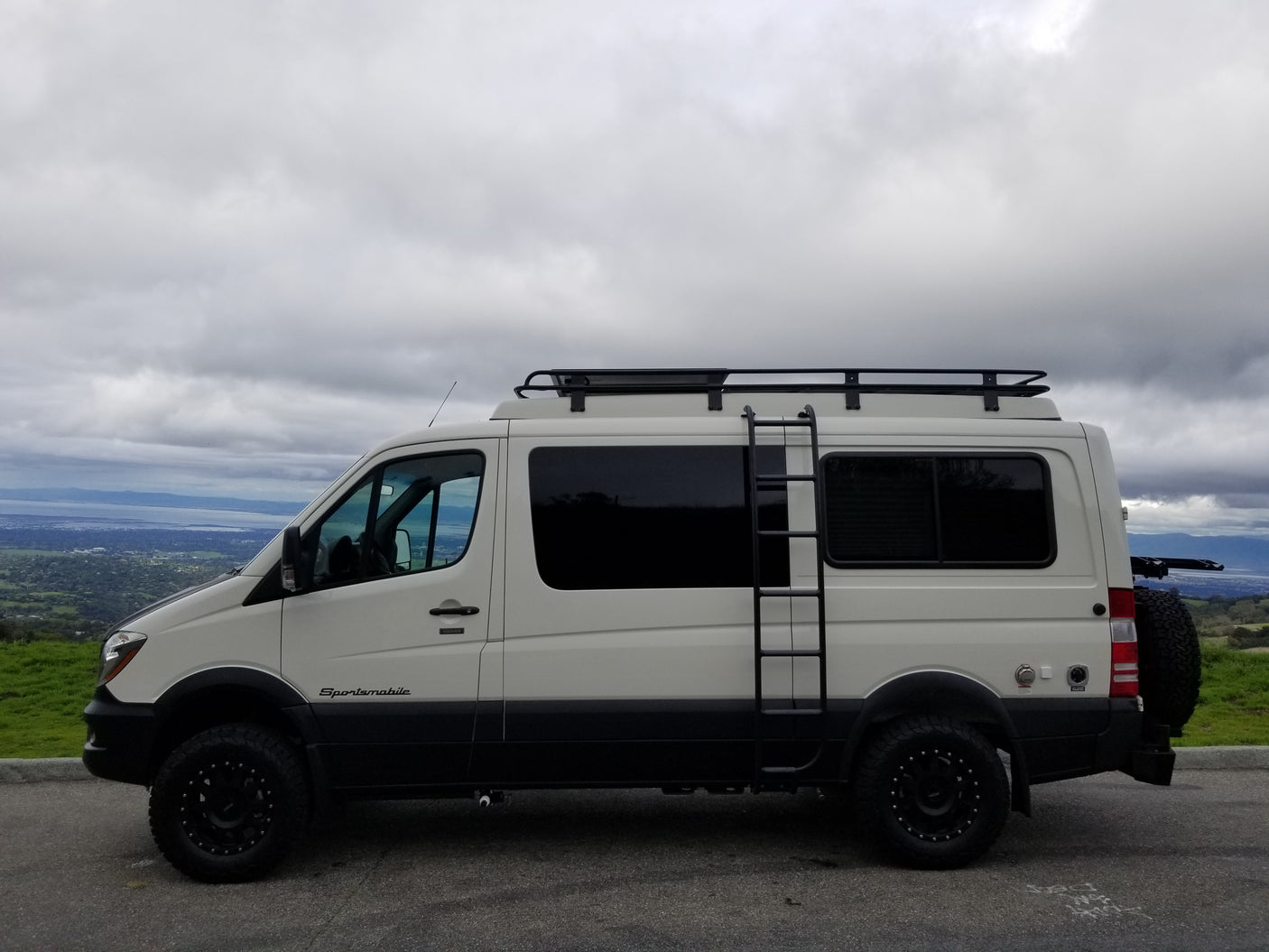 White Sportsmobile Sprinter camper van, an excellent rv rental for off-road adventures, parked with a mountain and bay view.
