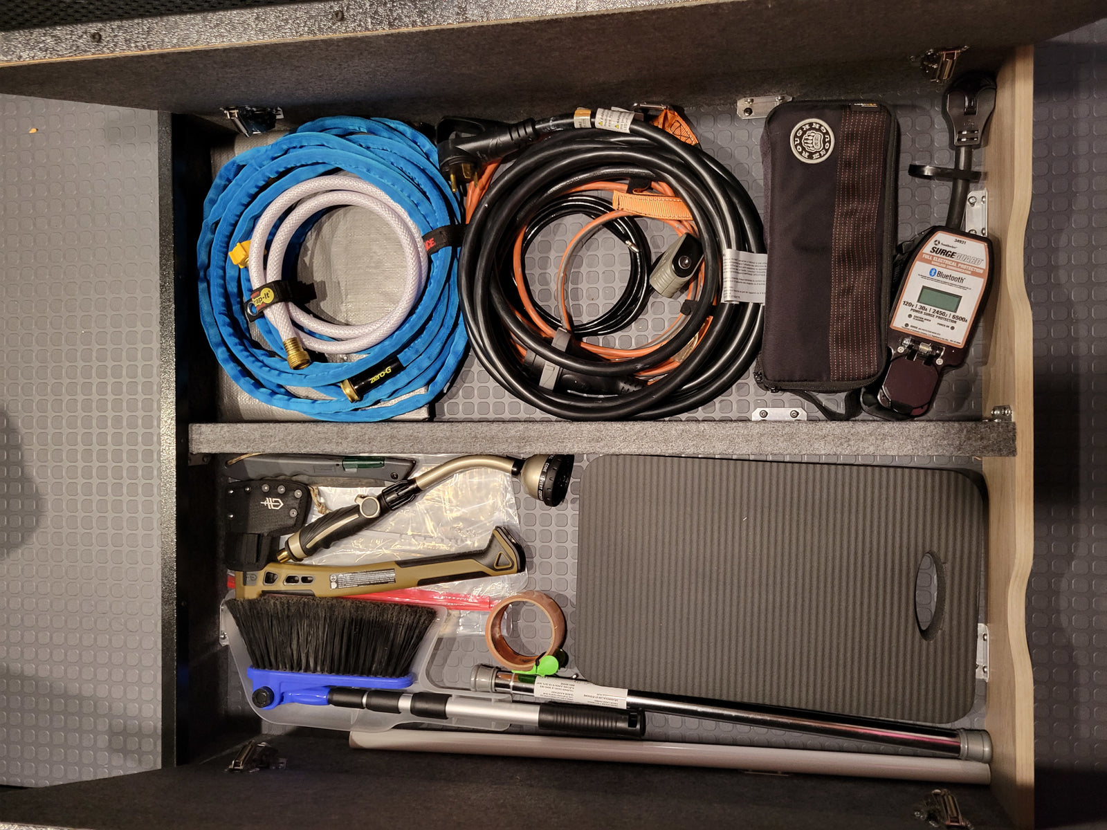 Organized RV rental compartment: water hose, power cords, Surge Guard electrical protection, and various tools neatly stored for travel.
