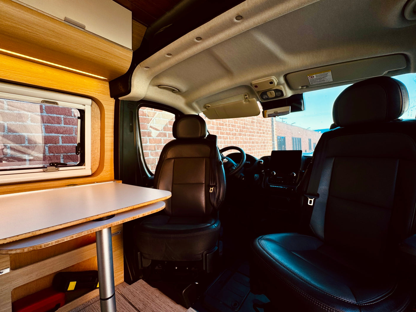Experience rv rental comfort: modern living area with wooden table & window, leading to the driver's cabin with leather seats.