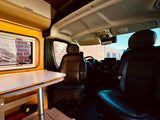 Experience rv rental comfort: modern living area with wooden table & window, leading to the driver's cabin with leather seats.