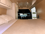 Cozy RV rental interior featuring a comfortable bed, overhead cabinets, warm wooden paneling, and the driver's cab.