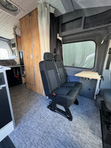 RV rental interior: custom camper van with two passenger seats, wooden cabinets, elevated bed, and gray flooring.
