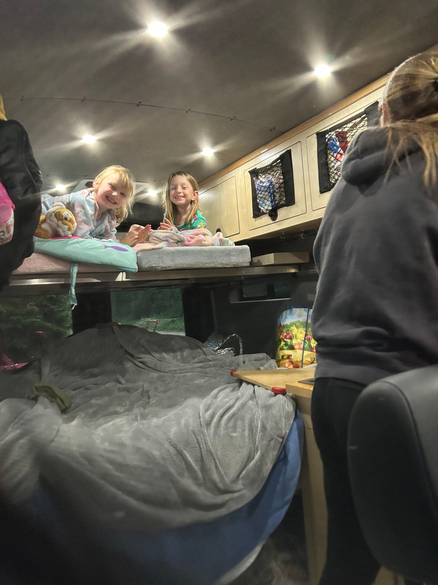 Two smiling girls are cozy on an RV rental bunk bed, ready for a family adventure. Inside a camper van at night.