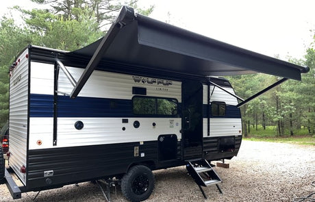 RV rental: a white & blue travel trailer with black awning extended, steps down, parked on gravel with pine trees.