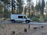 White camper van at campsite 20 amidst pine trees. Perfect RV rental for nature getaways.