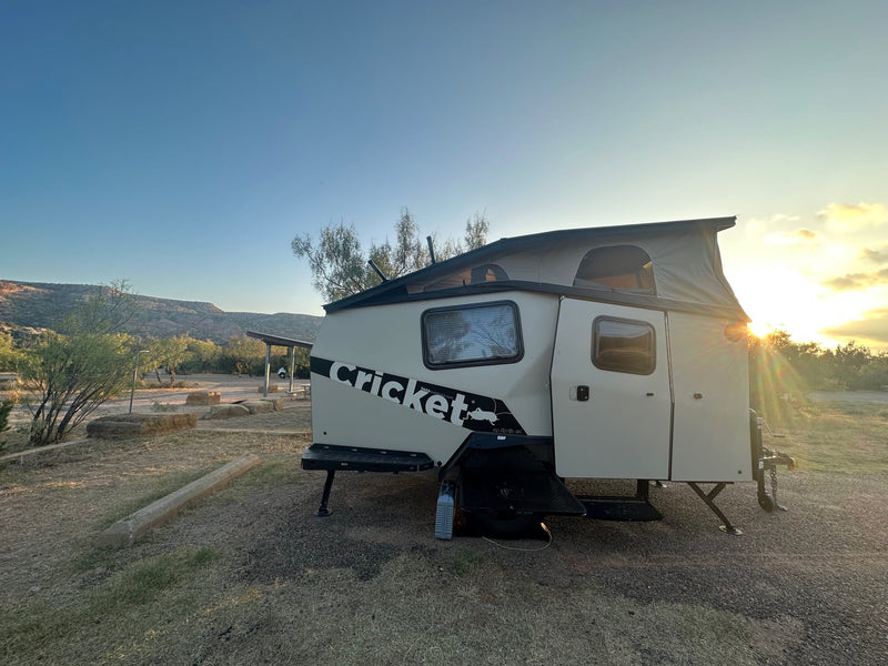 Light-colored Cricket camper (rv rental) with pop-up roof parked in nature. Golden sunlight pierces through trees at sunset.|An RV rental — photo 1